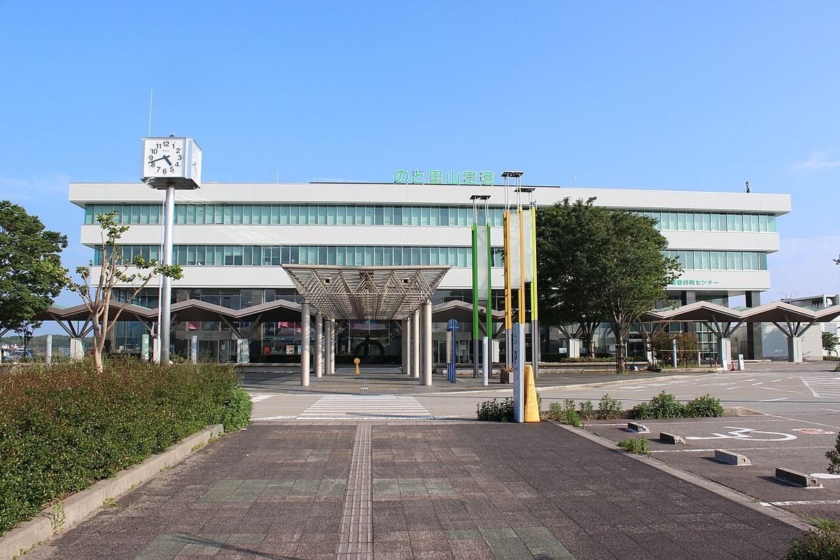 能登機場航廈正面,白色三層樓建築上掛著綠色的「のと里山空港」字樣,左前方立著一座四面時鐘的鐘塔,航廈前有雨遮與三根旗桿,前方的接送車道與停車格空無一車一人