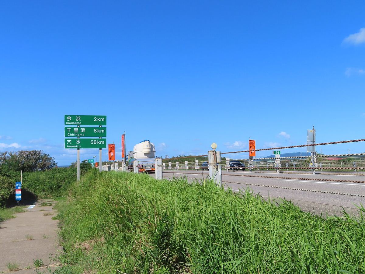 のと里山海道路旁的綠底距離指示牌,由上而下寫著今浜 2km、千里浜 8km、穴水 58km,牌子後方是正常通車的雙向車道,一輛槽車與幾輛轎車在行駛,天空晴朗無雲