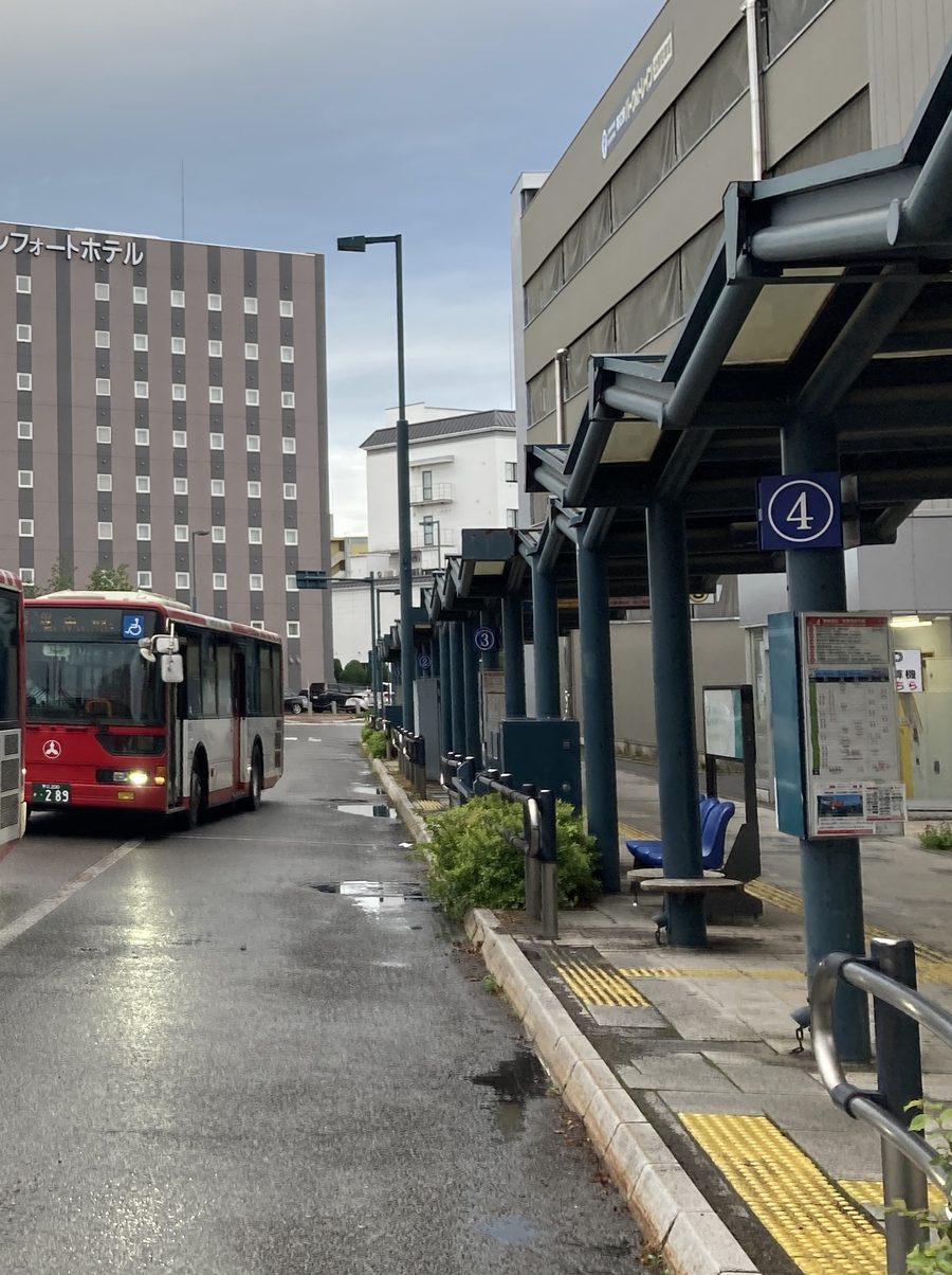 帶廣站巴士總站的候車月台:深藍色鋼柱撐著長雨棚,柱子上掛著藍底白字圓圈「4」號牌和時刻表看板,往後依序是 3 號、2 號;左邊車道上停著紅白色的拓殖巴士