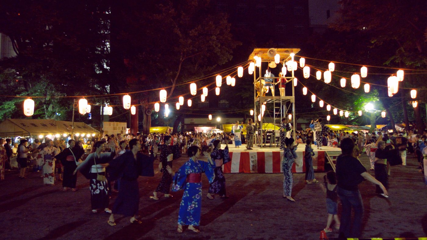 夜間盆踊會場：中央高台（櫓）掛滿紅白燈籠，太鼓手站在台上，穿浴衣的人群圍著高台繞成同心圓緩慢起舞