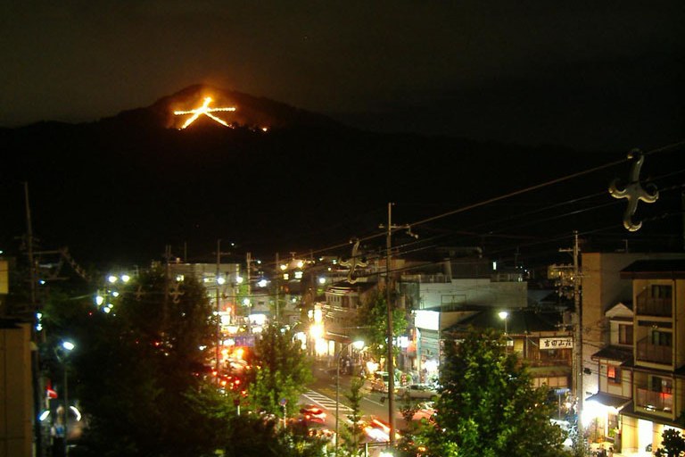 京都五山送り火的大文字：如意ヶ嶽山腹上由火床排成的巨大「大」字在夜空中燃燒，前景是京都市區屋頂與零星燈火