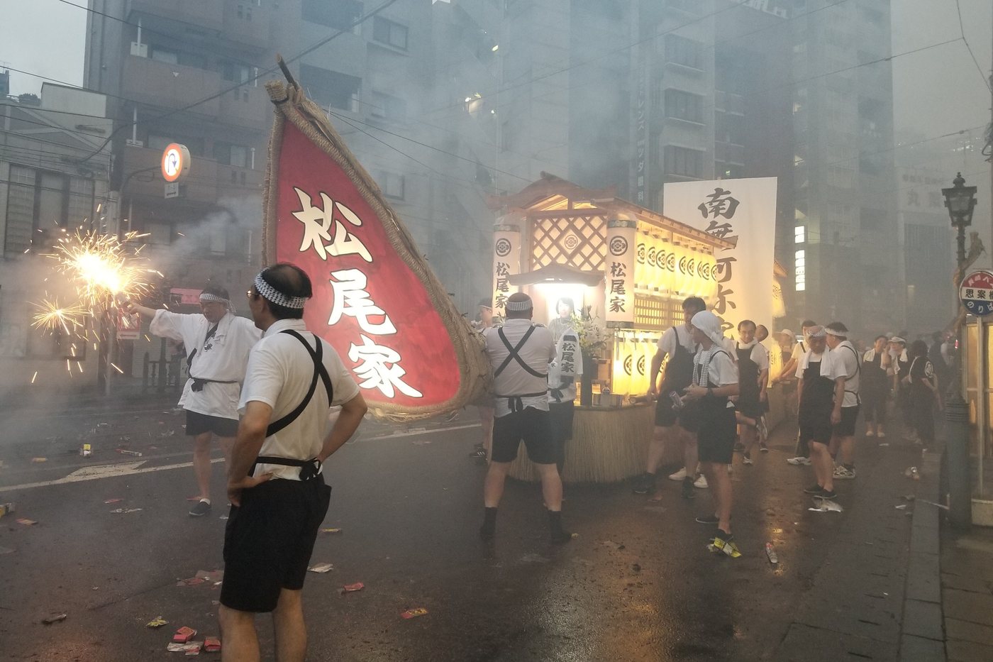 長崎精霊流し：家屬合力推著掛滿燈籠與紙花的木造精霊船走在夜間街道上，前方地面散落大量爆竹殘骸與白煙