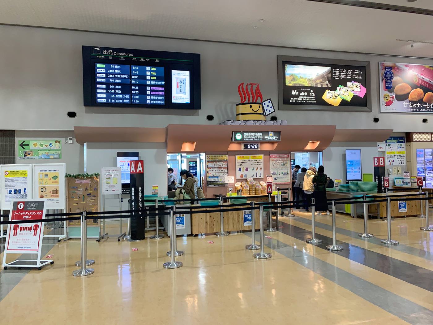 大分機場出發樓層,檢查場上方的深綠色看板寫著「国内線保安検査場 Domestic Security Check」,下方電子鐘顯示 9:28,左右標著 A、B、C 通道,牆上並排三塊告示板,中間那塊畫著外套與靴子的彩色圖示,檢查台前堆著印有 OITA 字樣的圓形木桶造型托盤