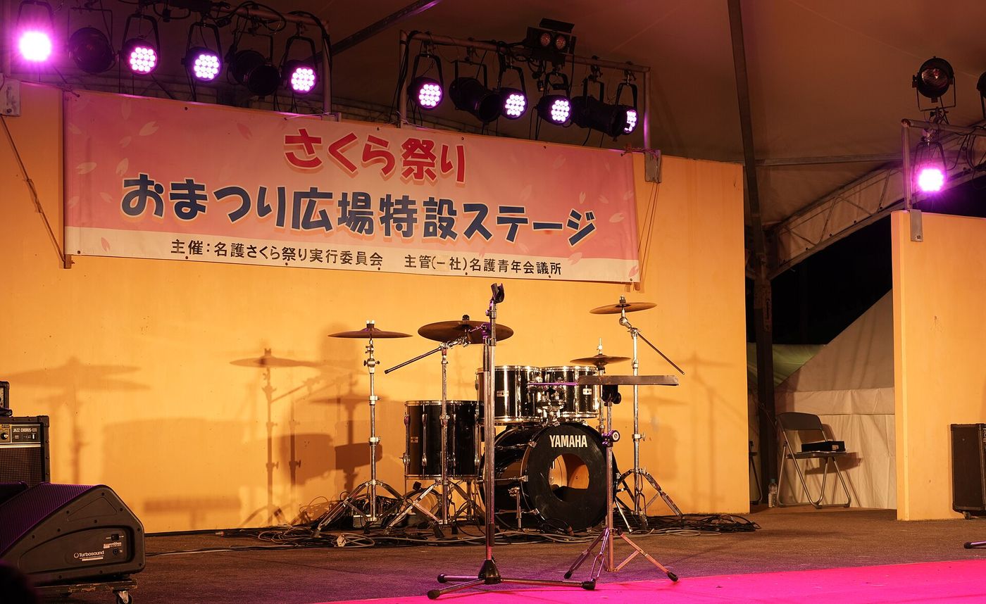 名護さくら祭り的戶外特設舞台:粉紅色布條寫著「さくら祭り おまつり広場特設ステージ」,舞台上架著一套爵士鼓與音箱,上方是一排粉紅色投射燈