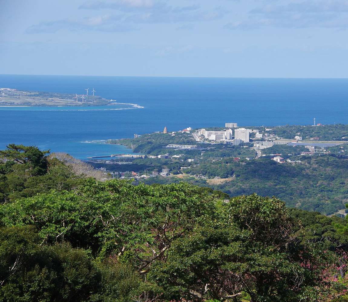 從八重岳往下看本部町:前景是常綠樹林,中景是市區與飯店建築,遠處是深藍色的海與一座架著風力發電機的岬角