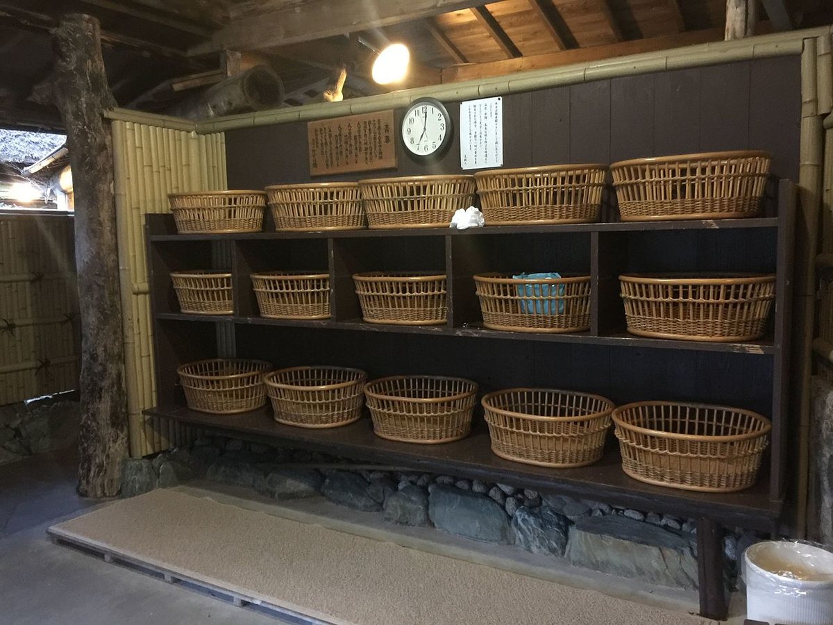 日本溫泉旅館的脱衣所一角:深色木架分三層擺著十五個藤編置衣籃,右下角有一個白色垃圾桶,籃架上方的木板牆掛著一塊直書的木製匾額、一個圓形掛鐘與一張直書的白紙告示,左側是竹編隔屏