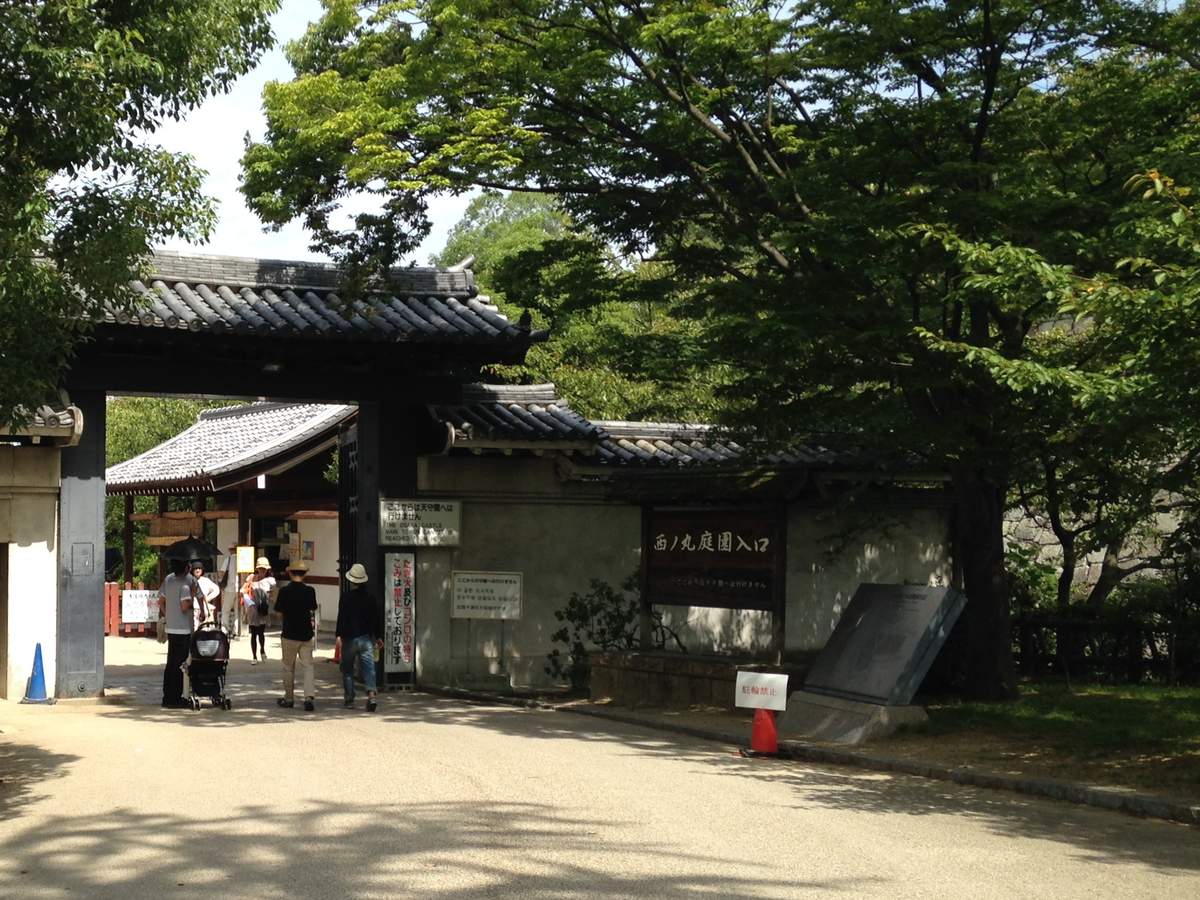 白天的西之丸庭園入口：瓦頂木門，右邊黑色木牌寫「西ノ丸庭園入口」，門柱旁白牌寫從這裡到不了天守閣，遊客背對鏡頭往裡走