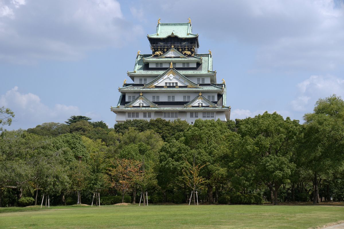 白天的大阪城西之丸庭園：前景一大片草坪，後面一排樹林，天守閣從樹林上方露出來，天空多雲
