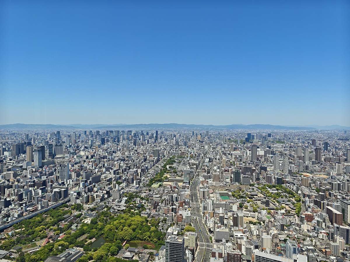 從阿倍野 HARUKAS 高處俯瞰大阪市區：整片低矮街區與高樓一直鋪到地平線，遠方一整排山稜的輪廓清晰可辨，天空是無雲的深藍色，畫面左下角是一片綠地與水池