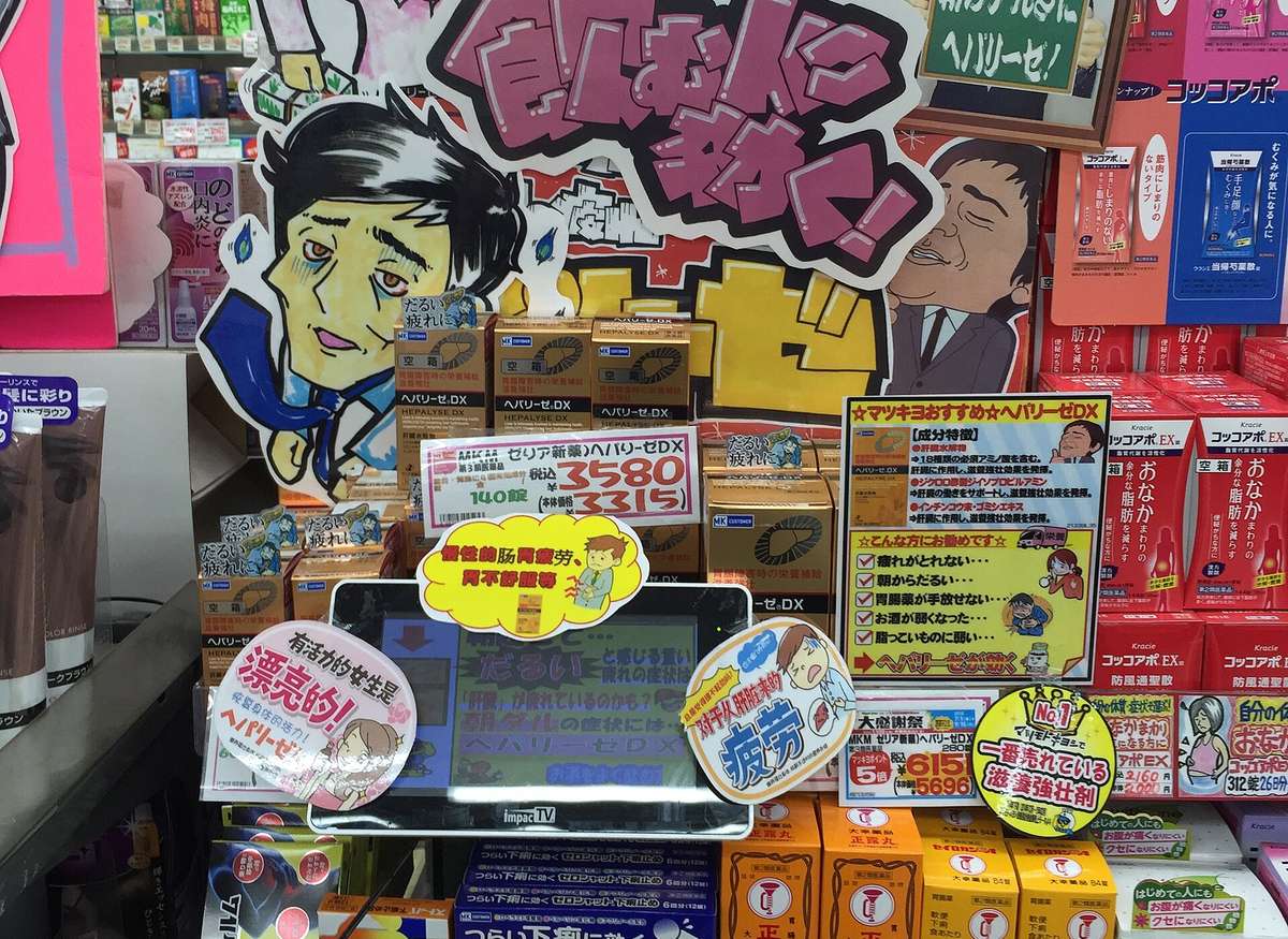 日本藥妝店的成藥區:層架上密密排著滋養強壯劑、胃腸藥與止瀉藥的紙盒,中間立著手繪的推薦看板與紅色手寫價格牌,好幾個展示盒的正面貼著寫有「空箱」的白色標籤。