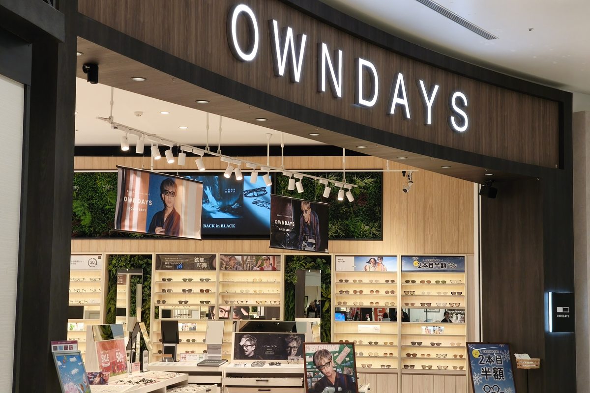日本購物中心內的 OWNDAYS 門市正面,發光店招下方是整面鏡框層架與中央的鏡框陳列台
