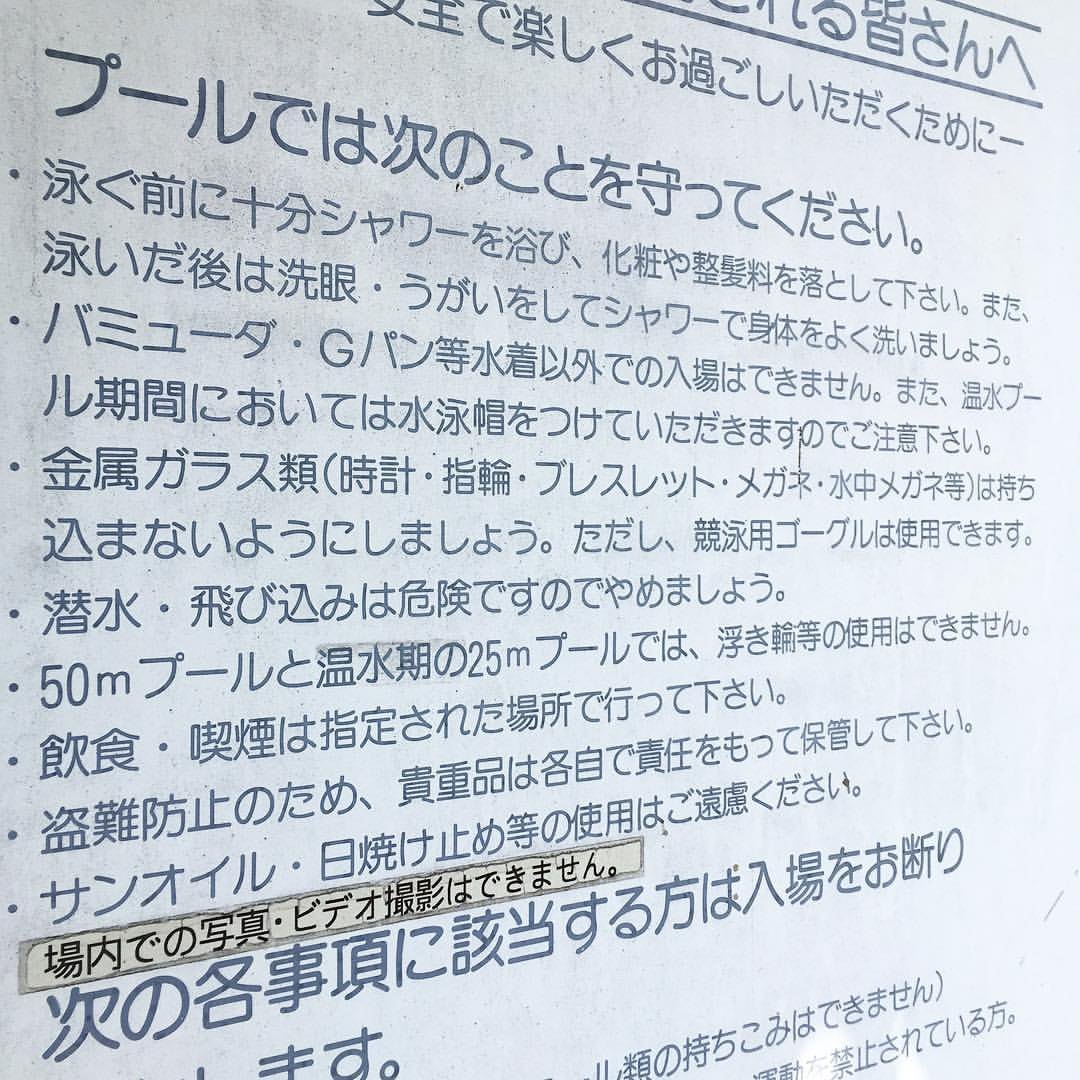 日本泳池的規則告示牌斜拍特寫:標題「プールでは次のことを守ってください。」下面一長串條列——游泳前先沖澡、バミューダ與牛仔褲等非泳衣不得入場、溫水池期間要戴泳帽、金屬玻璃類請勿帶入、禁止潛水與跳水、50 公尺池不可用浮圈、飲食與吸菸限指定場所、禁用防曬油;下方另貼一條「場内での写真・ビデオ撮影はできません。」,最底下是「次の各事項に該当する方は入場をお断りします。」
