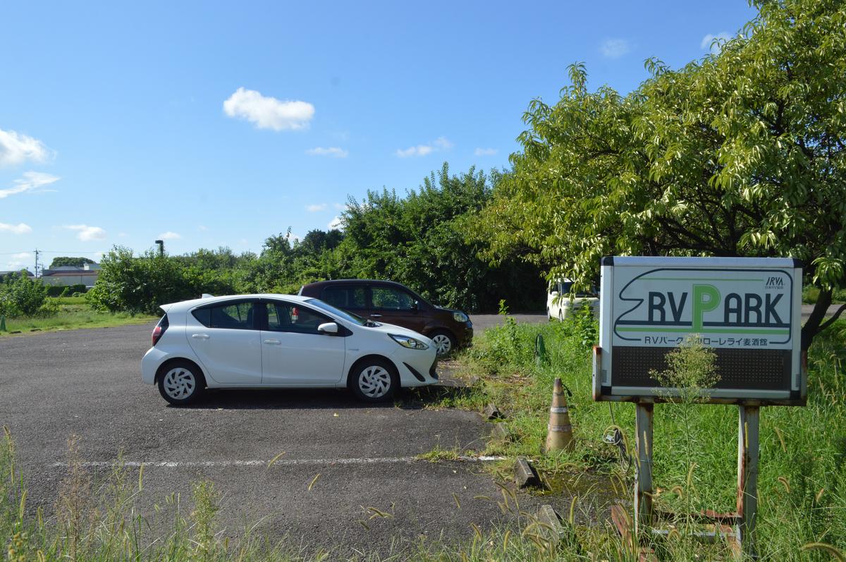 一塊寫著「RV PARK」與日本RV協会標記的白色認證看板立在草叢邊,旁邊的柏油空地停著一輛白色小客車與一輛暗紅色休旅車,後方是果樹與藍天。