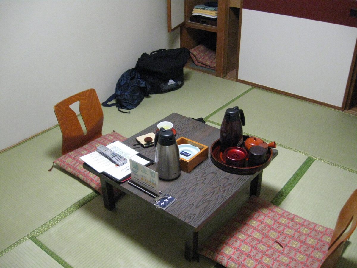 日本旅館的榻榻米客室,中央一張矮桌上擺著茶具、熱水瓶與遙控器,兩側鋪著座墊與無腳座椅,後方壁櫥開著、裡面疊著浴衣與備用座墊