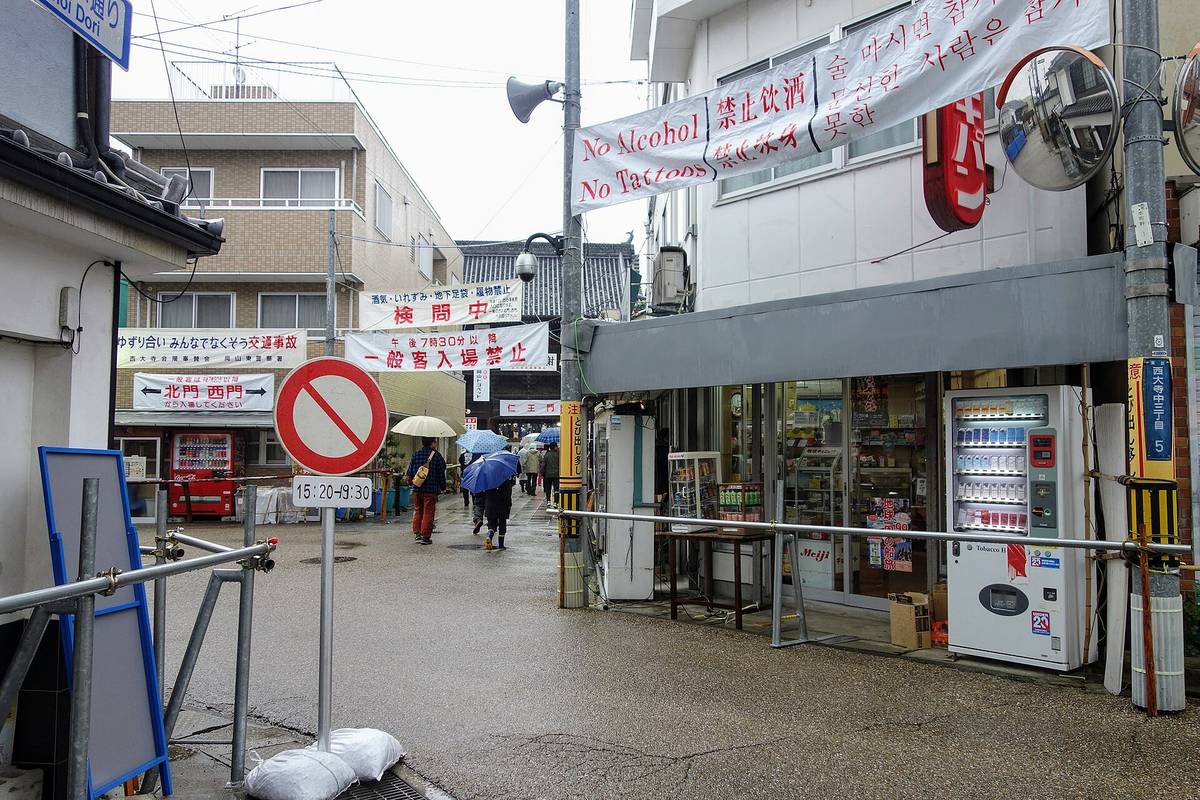 雨天的商店街街口,路面濕亮:橫掛在半空的白色布條印著「No Alcohol/No Tattoos」與中文「禁止饮酒/禁止纹身」及韓文;前方另一條紅字布條寫「酒気・いれずみ・地下足袋・履物禁止 検問中」,其下白底黑字寫「午後7時30分以降 一般客入場禁止」;左側看板寫「北門 西門から入場してください」,路中央立著一面禁止進入的圓形交通標誌,下方時間牌寫 15:20-19:30;街道深處有幾名撐傘的行人背影走遠
