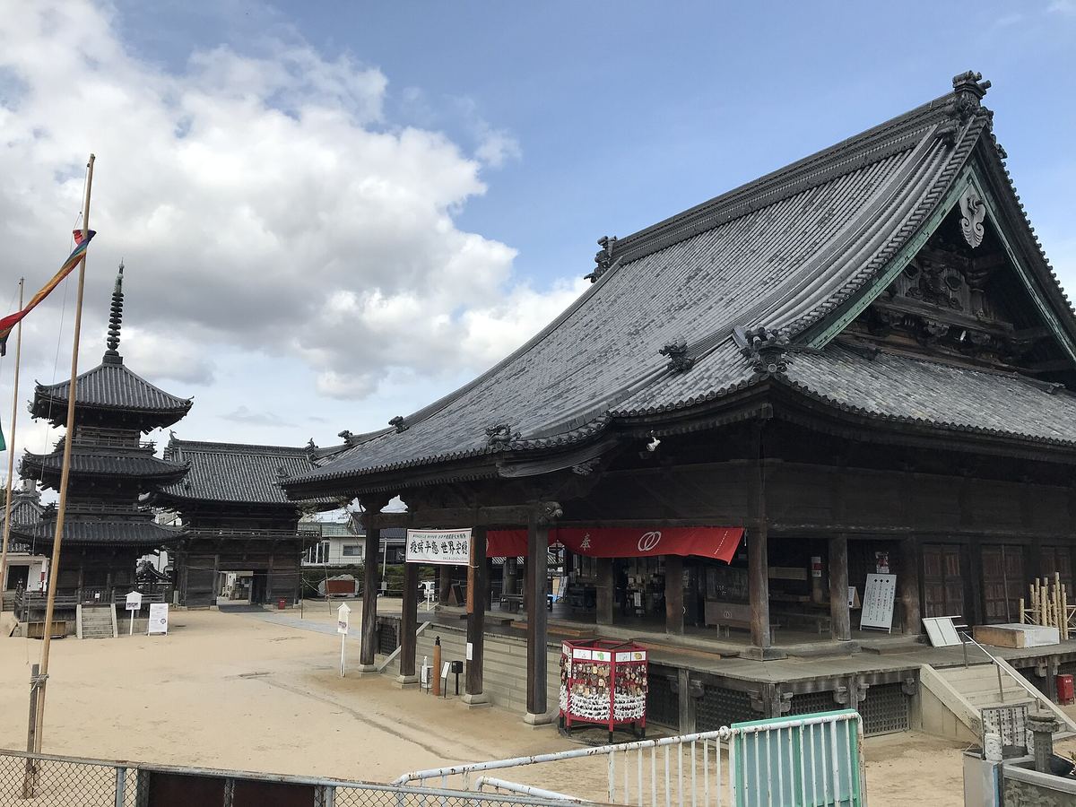 西大寺観音院的本堂近景:巨大的灰瓦屋頂與大幅翹起的簷角,正面木柱之間垂著一整條紅色布幕,柱前是抬高的木造平台與一具紅色的賽錢箱,庭中鋪著沙土,左後方是三重塔與寺務所,天空有積雲