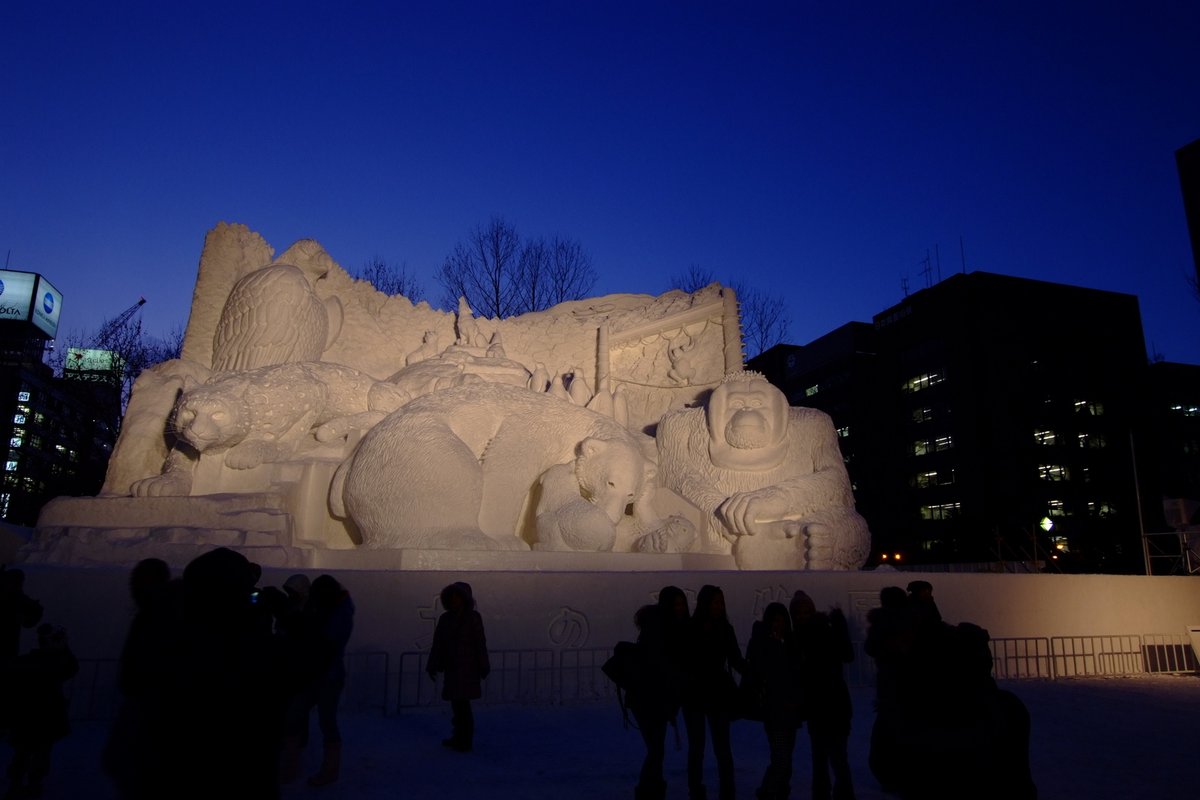 札幌雪祭大通會場 2010 年的一座大雪像,雕著雪豹、北極熊、紅毛猩猩、老鷹與企鵝,傍晚打燈,前方一排遊客的黑色剪影只到雪像底座高度