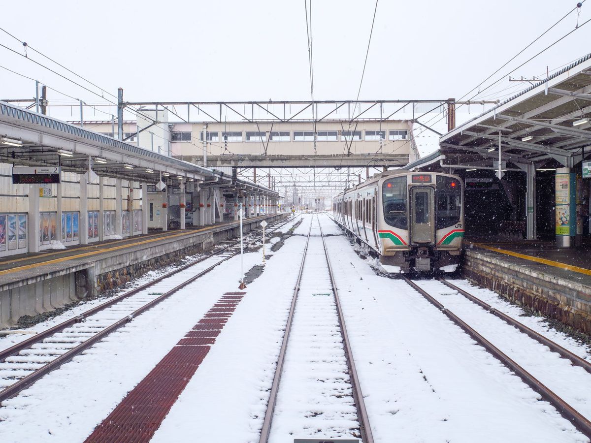 下雪中的會津若松站:月台之間的軌道積滿了雪,右側月台邊停著一列紅綠色塗裝、車頭有 JR 標誌的 E721 系電車,月台上沒有人