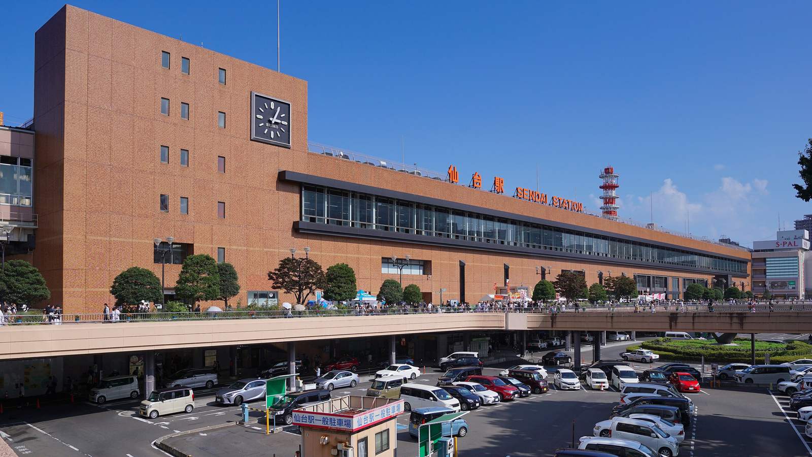 晴天的 JR 仙台站西口外觀:長條形褐色磚牆站房,左側方塔掛著大時鐘,屋頂上是紅字「仙台駅 SENDAI STATION」,站前是一層有許多行人的高架步行平台,平台下是停滿車的停車場與寫著「仙台駅一般駐車場」的收費亭,右後方是 S-PAL 百貨