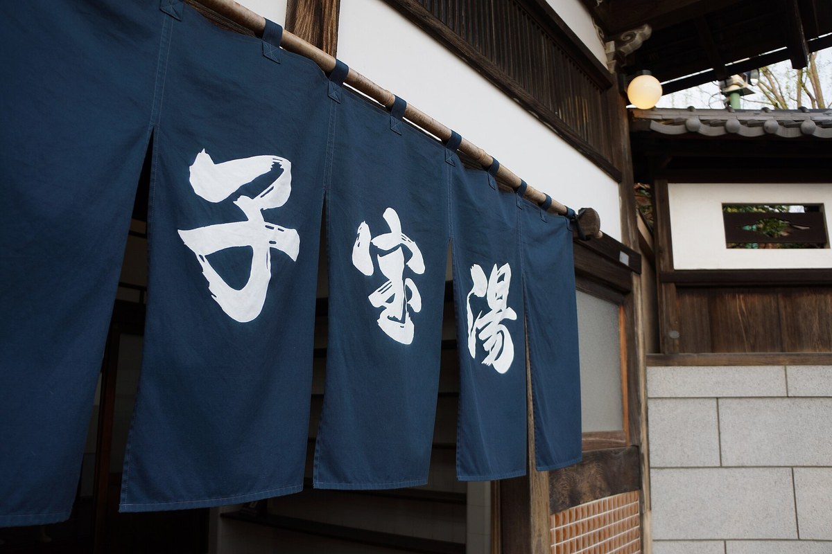 錢湯「子宝湯」入口的藍染暖簾特寫，深藍布面染著白色「子宝湯」三字，掛在竹竿上、上方是老式木造屋簷