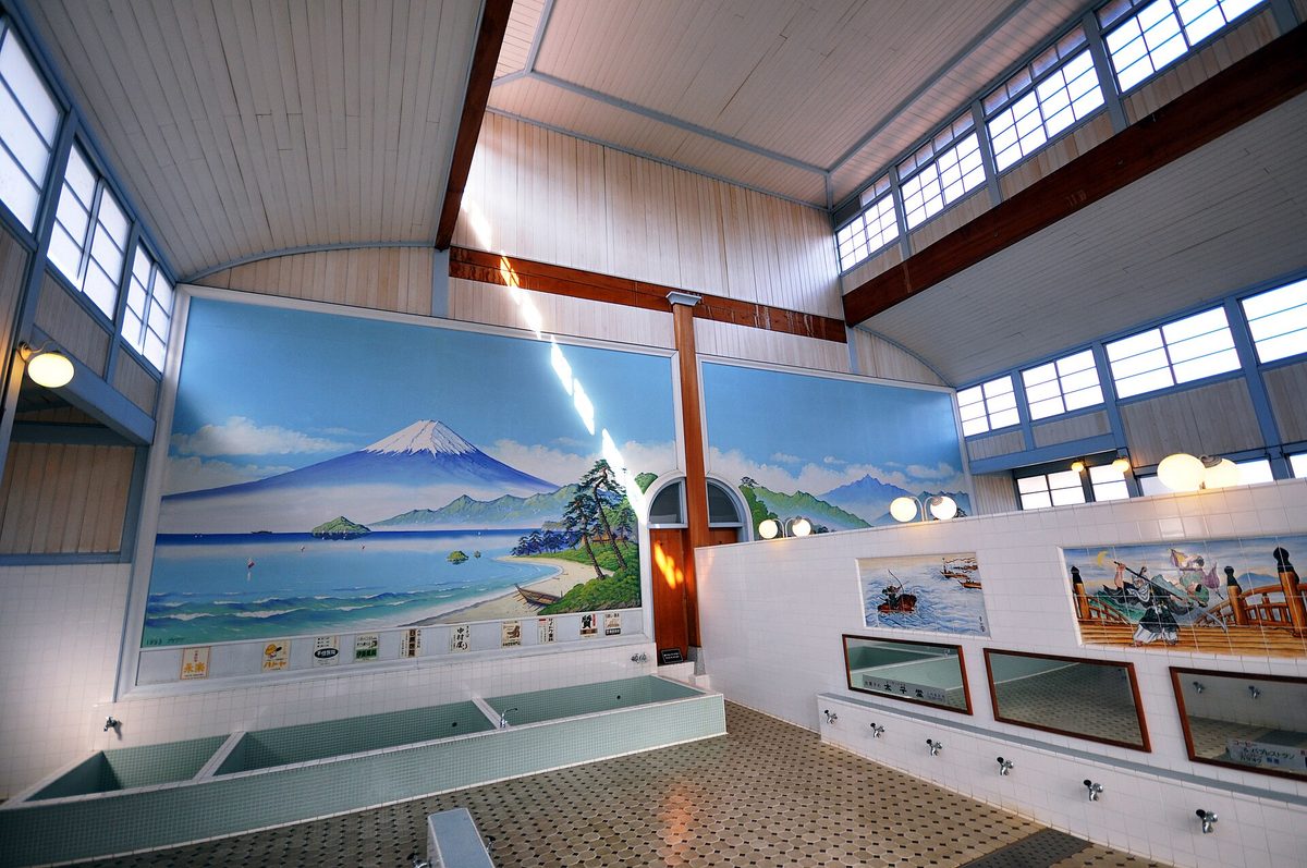 錢湯浴室全景：正面牆上是富士山與海岸的彩繪壁畫，左側是磁磚砌的長方形浴槽，右側牆邊一整排裝在矮牆上的水龍頭與鏡子，地面鋪著八角形磁磚