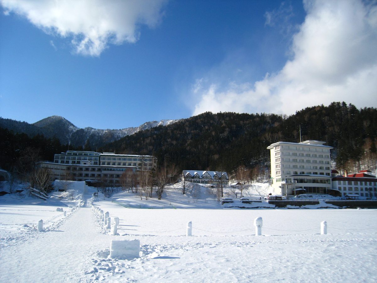 從結冰的然別湖湖面往岸上看:前景是冰上村的雪柱步道,岸邊右側是白色多層樓的ホテル風水,中間一棟半木造山形屋頂的小建築,左側是另一棟長形旅館,背後是覆雪的山