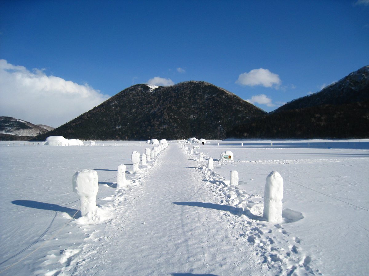然別湖冰上村的湖面步道:兩側立著一排雪柱,步道一路通到遠處湖面上的幾棟冰屋,背後是整片覆著樹林的天望山與藍天