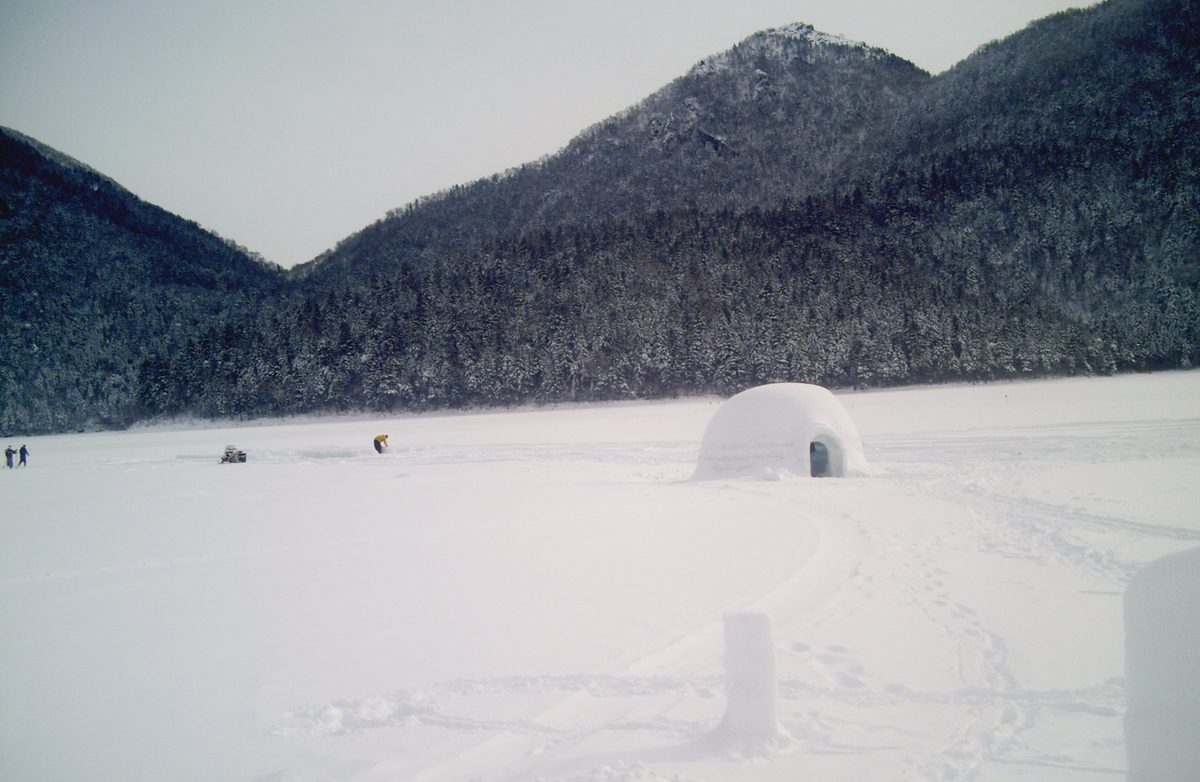 1 月下旬陰天的然別湖湖面,一大片雪地上只有一棟圓頂冰屋,遠處有幾個人和一台雪上車在湖上作業,背後是覆雪的針葉林山坡