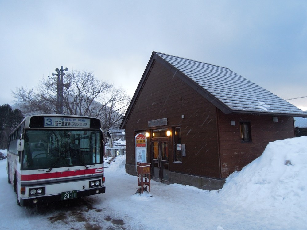 下雪的傍晚,北海道中央巴士「支笏湖」站:紅白塗裝的路線巴士停在積雪路邊,車頭顯示「3 千歳駅前経由 新千歳空港」,旁邊是咖啡色木造候車小屋和站牌,屋旁堆著雪