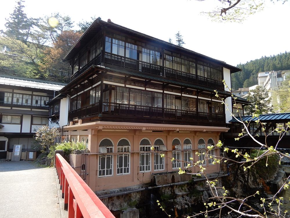 積善館本館正面：三層木造建築，二樓與三樓是整排木框玻璃窗配黑色欄杆，底層是粉橘色的石造基座、開著一排半圓拱窗；畫面左下角是慶雲橋的紅色欄杆，橋直接通到旅館前