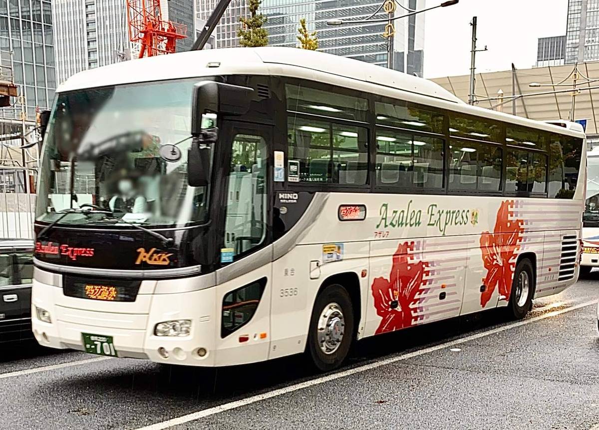 關越交通的高速巴士停在東京市區路邊：白色車身印著紅色杜鵑花圖案與 Azalea Express 字樣，車頭下方的橘色顯示器寫著「四万温泉」，背景是玻璃帷幕大樓與紅色工程吊車