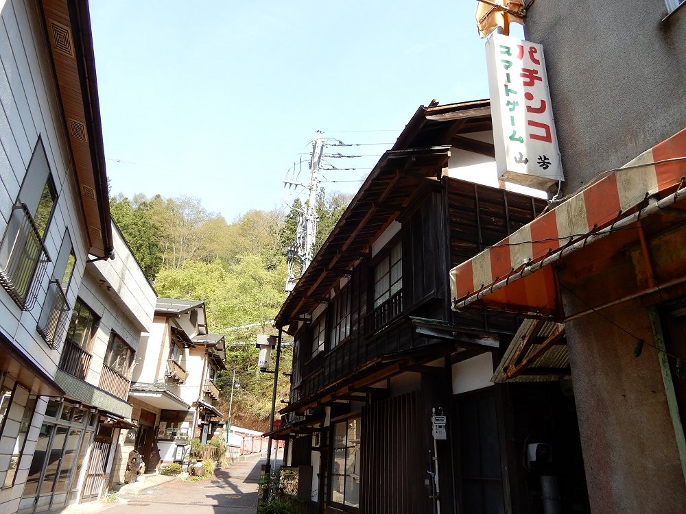 四万温泉落合通り的小巷：右邊是兩層樓的木造老屋，掛著「パチンコ」直式招牌和一片鏽掉的橘白條紋遮雨棚，木門全部關著，巷子往山坡上延伸，路上沒有人