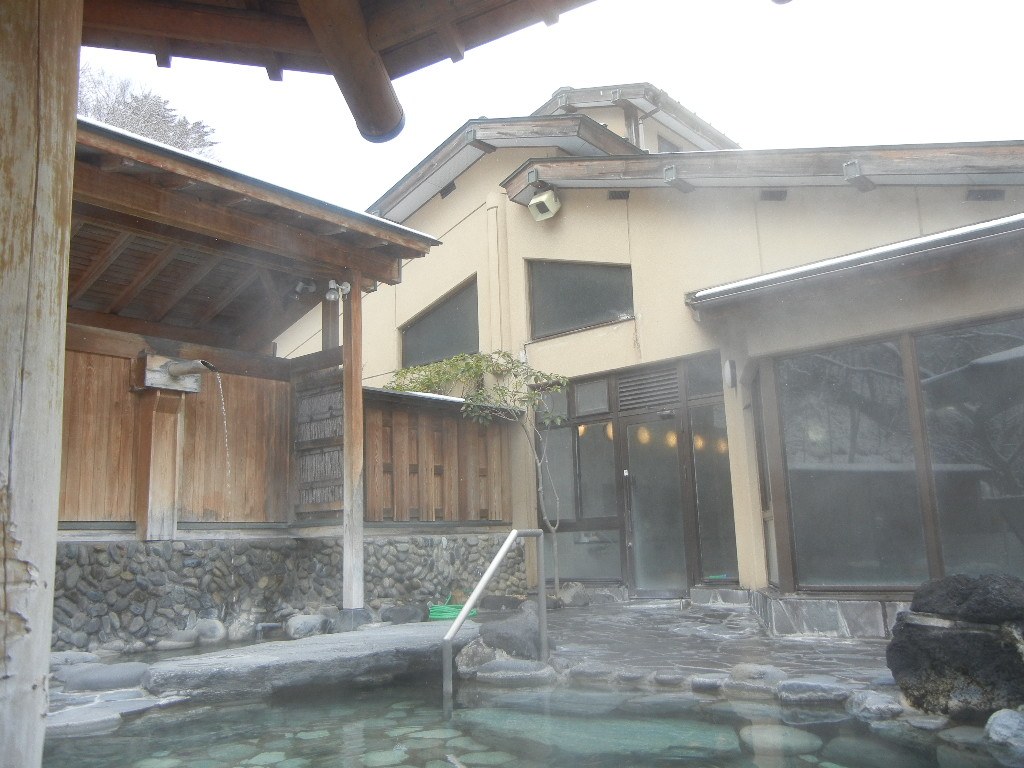 四万清流の湯的露天風呂：石頭砌成的浴池冒著白煙，左邊有一段木造屋頂和竹簾圍牆，一支木管把溫泉引進池子，後方是裝著大片落地窗的浴場建築