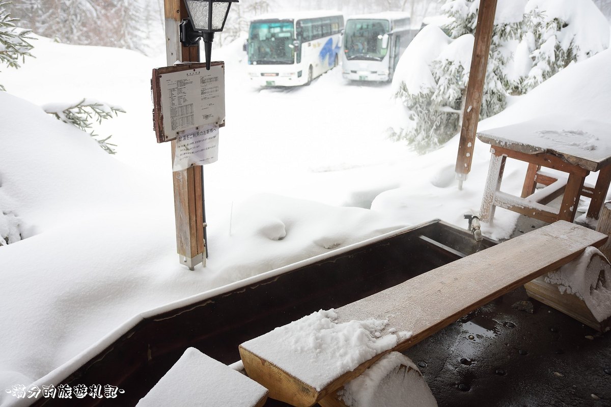 新穗高溫泉站前的戶外足湯,長條湯槽旁的木凳積著雪,柱上掛著溫泉分析書與足湯注意事項,後方雪地裡停著兩台濃飛巴士