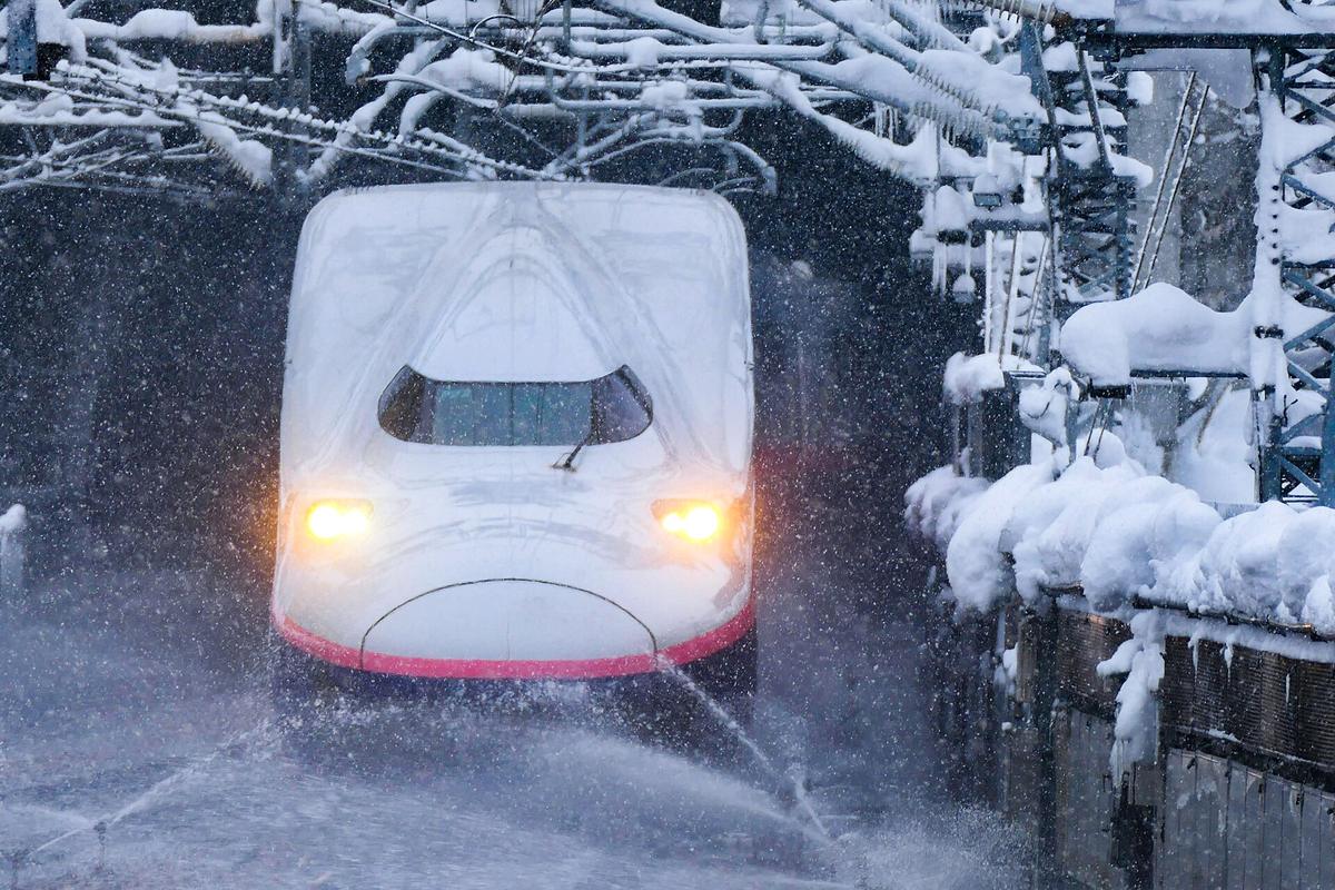 降雪中的上越新幹線列車正面特寫，車頭燈亮著駛入越後湯沢站，軌道旁的散水消雪噴水器正噴出水柱，架線支柱與圍籬上積著厚雪