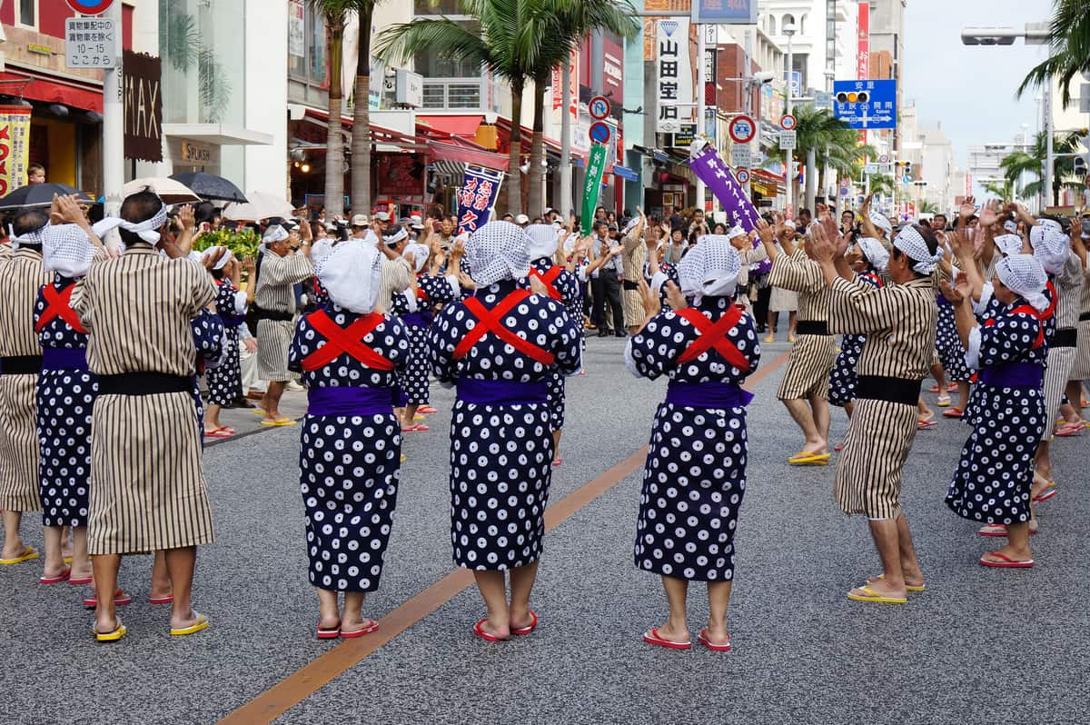 首里城祭期間的國際通:柏油路被封起來讓表演隊伍使用,前排是穿深藍底白色圓點浴衣、紫色腰帶、背後綁紅色交叉襷、頭上包白布的舞者,後排是穿米褐色直條紋和服的舞者,全體舉手拍掌,路旁店家騎樓下站滿撐傘的觀眾,背景可見棕櫚樹與「山田宝石」等招牌