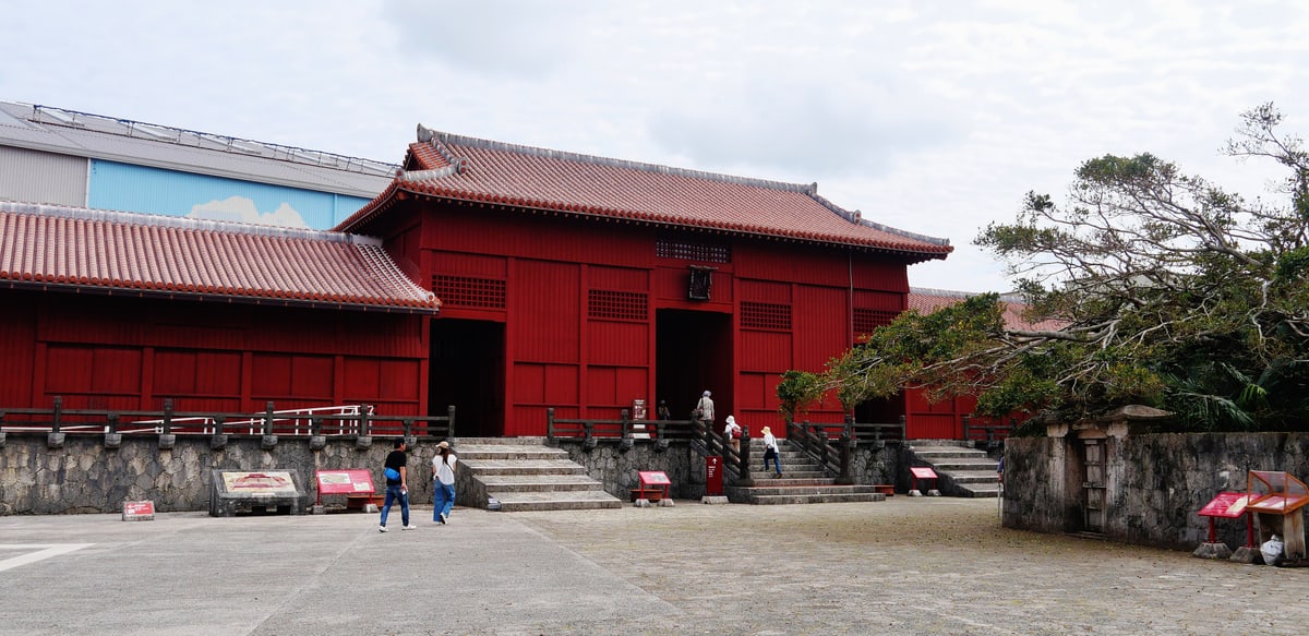 首里城下之御庭:奉神門前一整片灰色鋪石廣場,廣場上散放著幾面解說牌與長凳,正面是朱紅色木造的奉神門與兩側低矮的紅色圍屋,門上方掛著一塊黑色匾額,右側有一棵大樹,畫面左後方露出一座白色與藍色的大型工事用構造物