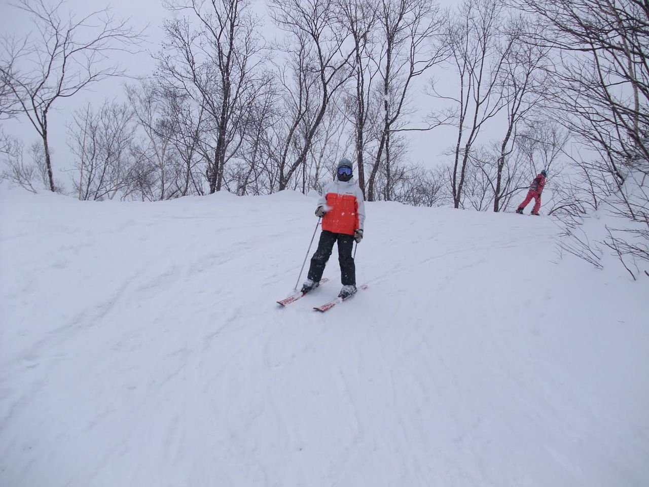 滑雪者正面全身照，戴淺灰色安全帽與藍色鏡片護目鏡、脖圍遮住口鼻，穿白底橘色胸片雪衣、黑色雪褲、淺色手套，雙手持雪杖踩著紅白雪板
