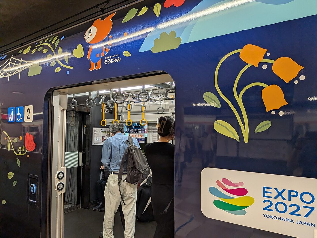 相鉄列車深藍色車身上的彩繪:橘色鐘形花朵、綠葉、紅花與蜘蛛網圖案,車門左上方印著標註「そうにゃん」的橘色貓角色,右下角一塊白底圓角牌貼著 EXPO 2027 YOKOHAMA JAPAN 彩色標誌,車門開啟中兩名乘客正要上車