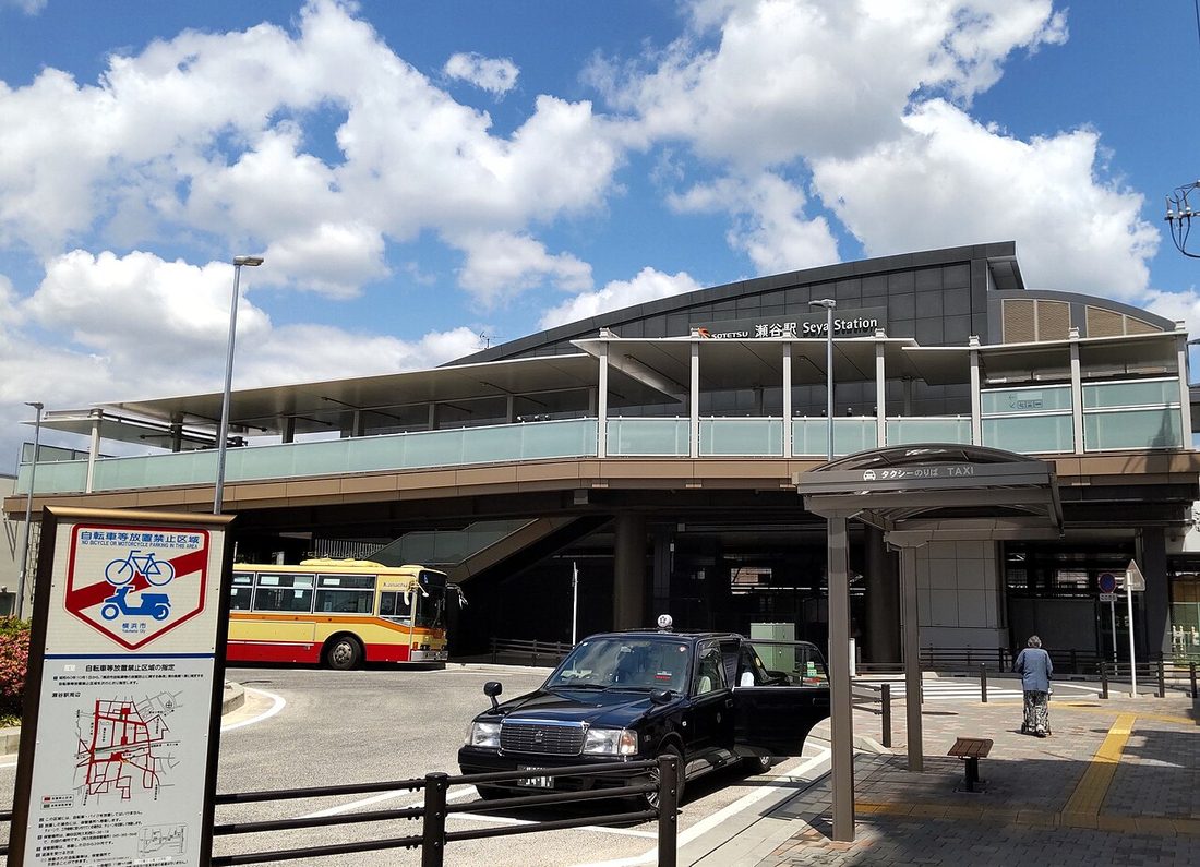 相鉄本線瀬谷駅高架站體外觀,站牆上掛著「SOTETSU 瀬谷駅 Seya Station」看板,站前有計程車招呼站遮棚、一輛黑色計程車與一輛黃紅配色的路線巴士,前景立著橫濱市的自行車禁止停放告示牌
