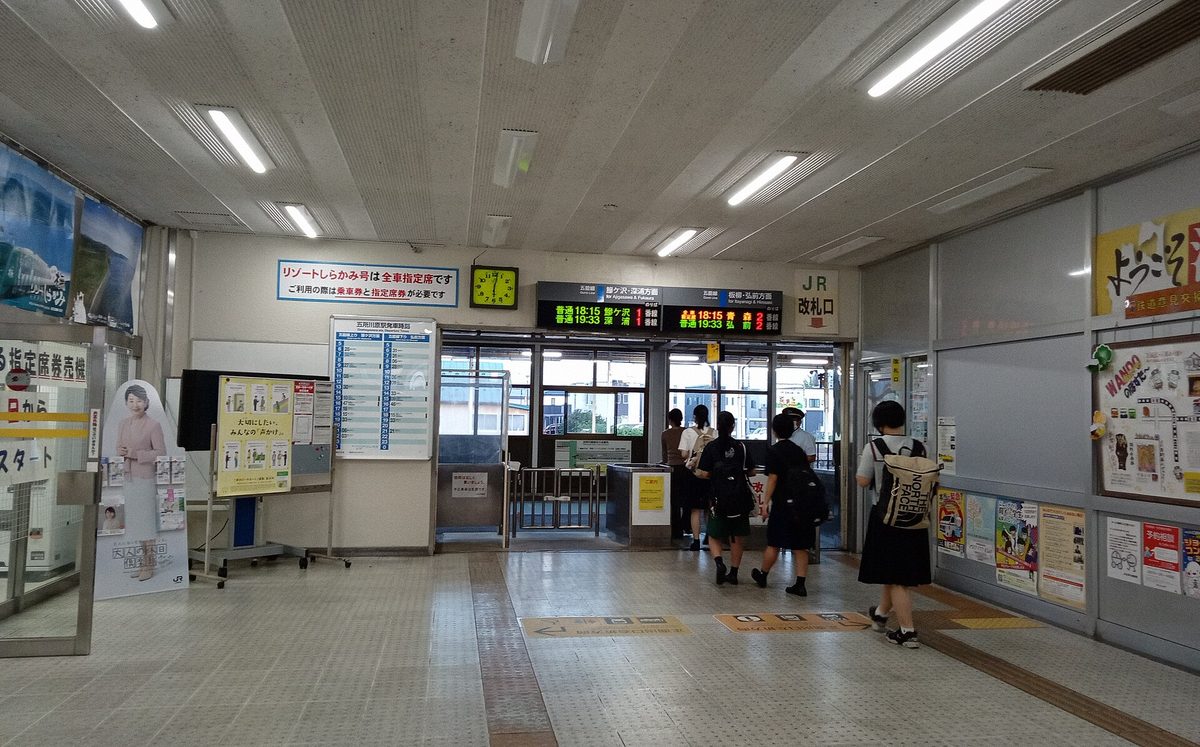 JR 五所川原站改札口，站員站在閘口旁逐一驗票，數名旅客背對鏡頭排隊通過，上方發車看板顯示兩班列車時刻