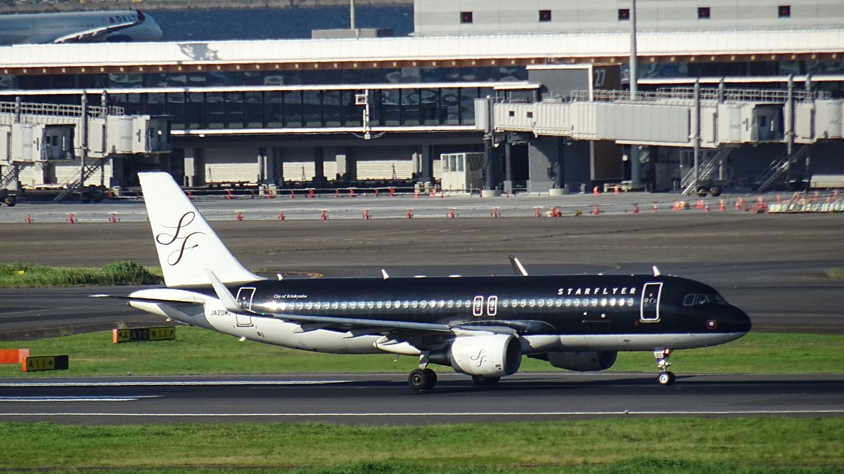 全黑塗裝的星悅航空 A320 在羽田機場滑行道上,前機身白字 STARFLYER、尾翼白底黑色 SF 花體字,機尾下方註冊編號 JA20MC,背景是航廈空橋