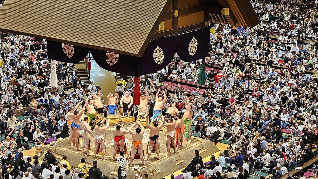 土俵入り儀式：十幾名力士穿著彩色化妝廻し在土俵邊緣站成一圈並舉起雙手，土俵上方吊著深紫色水引幕與金色紋樣，四周桝席與看台坐滿觀眾