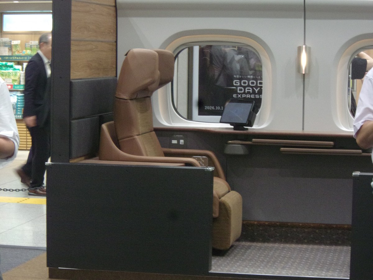 車站大廳裡的 Supreme Class Cabin 全尺寸模型：白色車身開著兩扇圓角車窗，室內是一張高背棕色皮革座椅與後方一張較低的座椅，窗下有木質檯面與兩根扶手桿，旁邊立著一台小平板，車窗內貼著寫有「GOOD DAY EXPRESS 2026.10.1」的黑色海報，左邊可以看到車站商店與走過的路人