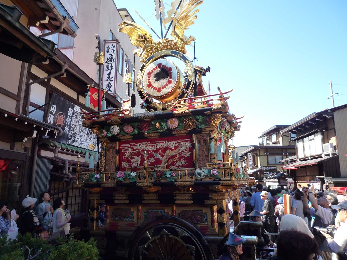 秋の八幡祭中停在高山老街上的屋台,金鳳凰與精緻雕刻在陽光下閃亮,兩側是圍觀人群
