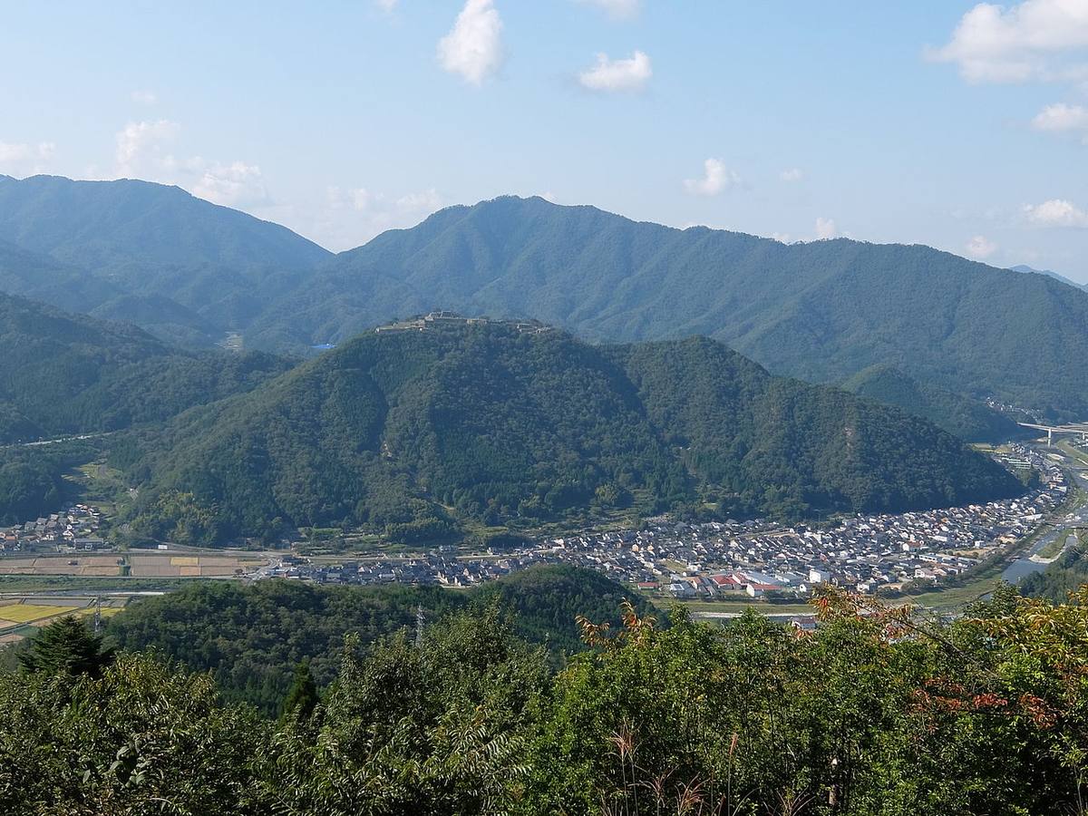 從立雲峡側越過谷地望向竹田城跡:城跡的石垣清楚地橫在對面山頂,山腳下是竹田城下町的住宅與稻田,右側是圓山川與國道