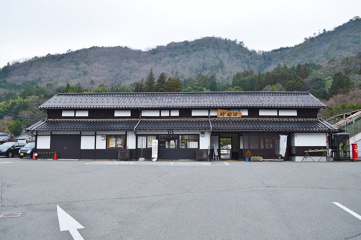JR 竹田駅站舍外觀:黑白相間的和風建築,屋簷下掛著木質「竹田駅」看板,站前是空曠的廣場,後方就是竹田城跡所在的山