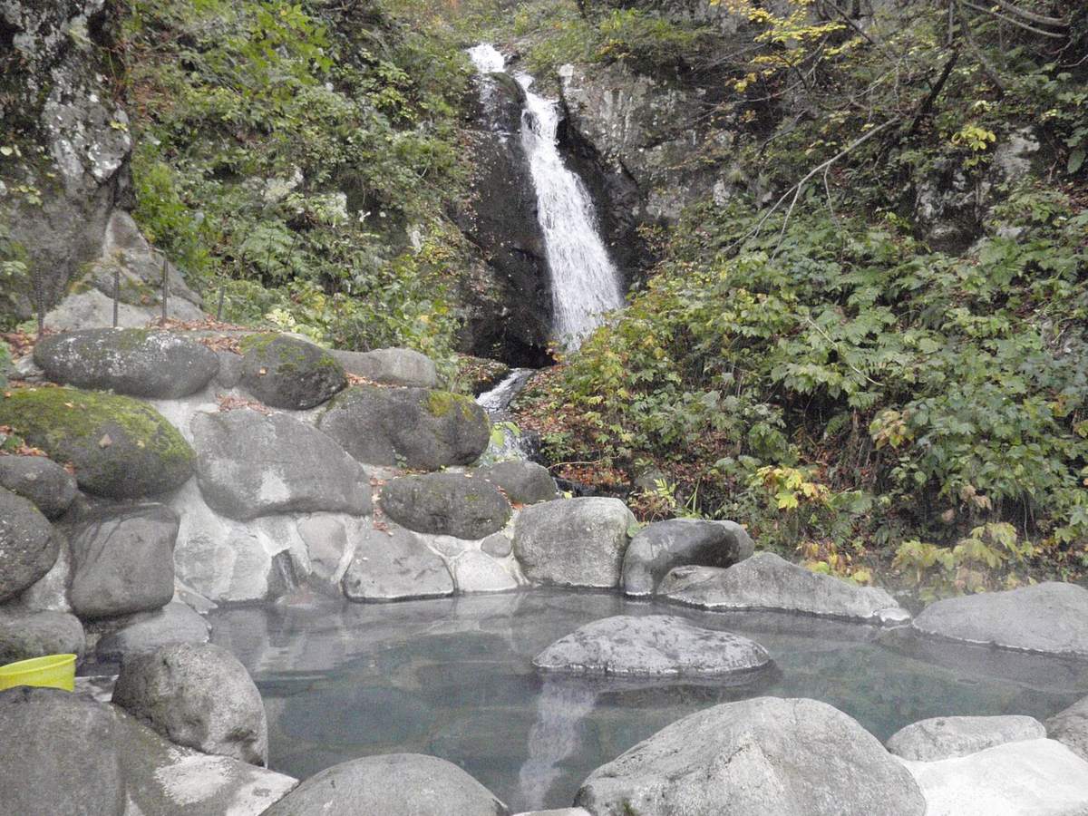 山中的岩砌露天溫泉池,水面冒著白煙、池中央露出一塊大石,後方岩壁掛著一道細長瀑布,周圍是綠色灌木與零星枯葉,左下角放著一個黃色塑膠桶