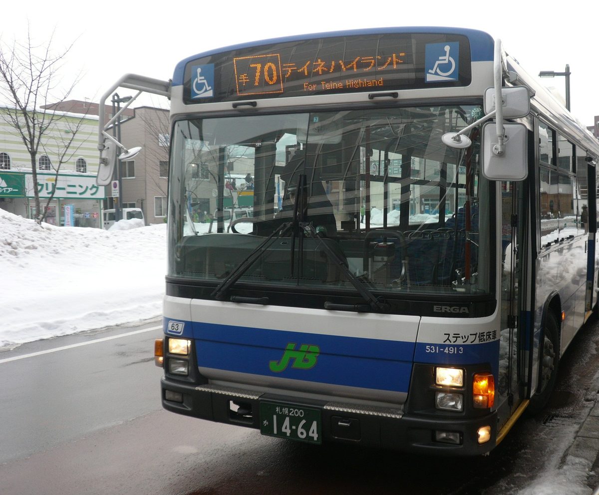 一輛白藍兩色的 JR 北海道巴士停在積雪的候車亭旁,車頭 LED 顯示器以橘字寫著「手70 テイネハイランド / For Teine Highland」,車身下緣印著綠色 JHB 標誌與「ステップ低床車」,路旁堆著剷起的雪堆