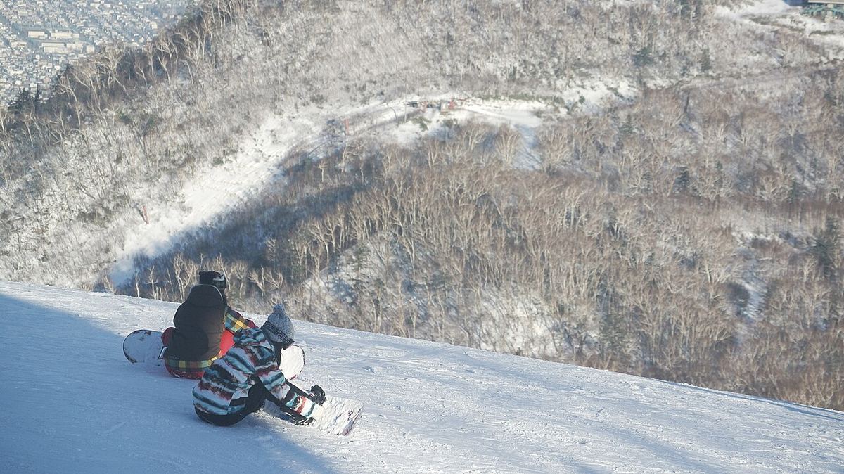 サッポロテイネ 的壓雪滑道邊坡,兩名單板客背對鏡頭坐在雪面上,坡下是落葉林谷地,左上角可見札幌市區的密集樓房